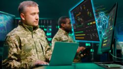 Military officer in camouflage standing in server room with laptop computer and checking data on monitors.